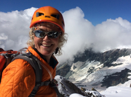 A person with curly blonde hair wearing an orange helmet, orange jacket, and sunglasses smiles at the camera. Equipped with a backpack, they stand on a snowy mountain during their hiking tour, with clouds and rugged peaks in the background.