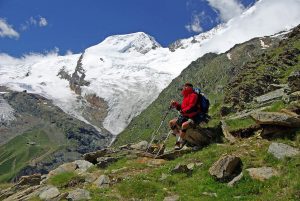 A person wearing a red jacket and carrying a backpack is sitting on a rock, using trekking poles while resting during their hiking tour. They are surrounded by rugged terrain and lush greenery, with a partially snow-covered mountain peak in the background under a clear blue sky.