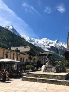 A sunny streetscape in a mountain village with snow-capped peaks in the background. People are sitting at outdoor tables under umbrellas near a yellow building, likely deciding on their next hiking tour. In the foreground, there is a stone statue on a pedestal. Blue sky with a few wispy clouds.