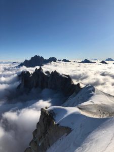 A breathtaking view of a snow-covered mountain peak rising above a sea of clouds, with rugged mountain ranges in the background under a clear blue sky. The scene exudes tranquility and majesty, perfect for a memorable hiking tour that highlights the harsh beauty of the high-altitude landscape.