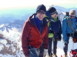 Three climbers, dressed in winter gear, stand on a snowy mountain peak. One of them, in a red jacket, is smiling at the camera while the other two, wearing blue jackets, look off towards the mountains. Snow-covered peaks and a clear sky are visible in the background—capturing the essence of their hiking tour.