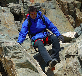 A person wearing a blue jacket, black pants, and red climbing harness navigates through rocky terrain on a hiking tour. The individual is equipped with a backpack and sunglasses, appearing focused on maintaining balance and grip on the uneven, rugged surface.