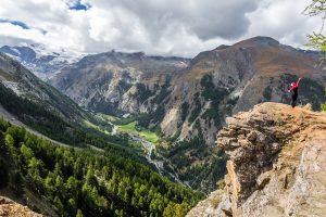 A person in a red jacket stands on the edge of a cliff overlooking a vast valley filled with dense green forests and rugged mountains under a cloudy sky. A village is visible in the valley below, surrounded by fields and trees, marking the perfect end to an invigorating hiking tour.