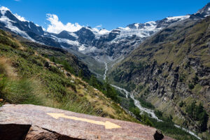 A mountainous landscape with snow-capped peaks beneath a clear blue sky. Rocky slopes and green vegetation cover the valley. In the foreground, a flat rock with a yellow painted arrow points towards the valley, indicating the trail direction for an adventurous hiking tour.