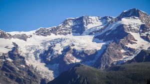 A majestic mountain range with snow-capped peaks against a clear blue sky. The rugged terrain features a combination of rocky cliffs and lush green slopes at the base, with patches of snow and glacial ice adorning the higher elevations, making it an ideal destination for a breathtaking hiking tour.