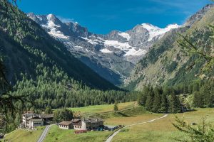 A picturesque mountain village nestled in a lush green valley, surrounded by towering snow-capped peaks and dense forests under a clear blue sky. A winding road leads to the village, which offers delightful rustic houses and buildings—an ideal starting point for an unforgettable hiking tour.