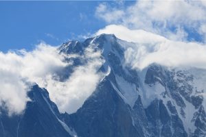 Snow-capped mountain peaks partially enveloped by wispy clouds under a bright blue sky. The rugged terrain of the mountains, perfect for a memorable hiking tour, is highlighted by shadows and sunlight, creating a dramatic and picturesque scene.