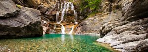 A serene waterfall cascades down tiered rock formations into a clear turquoise pool surrounded by rugged cliffs. The water is so transparent that smooth stones at the bottom are visible. Nearby, lush green foliage adds a touch of vibrancy to the scene, making it an ideal spot for a hiking tour.