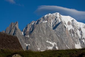 A breathtaking landscape showcasing a snow-capped mountain peak under a clear blue sky. In the foreground, a slightly visible wooden rooftop and grassy area invite thoughts of a hiking tour. The mountain is a mix of rugged gray rock and snow, with a unique cloud formation hovering above.