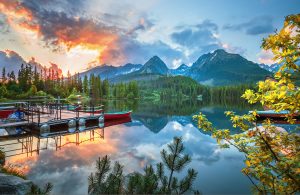 A serene mountain landscape at sunset. Tall conifer trees and lush greenery surround a calm lake with clear reflections of the sky and mountains. A dock with red boats is on the left, awaiting adventurous spirits returning from their hiking tour as the sky glows with vibrant colors of orange and blue.