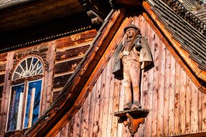 A wooden sculpture of a man in traditional attire is mounted on the exterior of a wooden building, often admired by those on a hiking tour. The building features an arched window with a sunburst design above it and intricate woodwork along its facade and roofline.