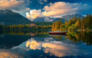 A serene lake reflects a vibrant sky, surrounding mountains, and lush forests. A small red and blue boat floats on the calm water. In the background, an A-frame building is nestled among the trees, with snow-capped peaks rising behind it—a perfect spot to start a hiking tour.