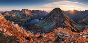 A breathtaking mountain landscape at sunrise with rocky peaks, a serene lake nestled between the mountains, and a mix of autumn colors in the foreground. The rising sun casts a golden glow over the scene, creating a warm, tranquil atmosphere—perfect for an inspiring hiking tour.