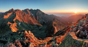 A panoramic view of a mountainous landscape at sunrise. The sun casts a warm, golden light on the rugged peaks and rocky terrain. Small patches of snow are visible among the rocks. Perfect for a hiking tour, the clear sky shows a gradient from deep blue to orange near the horizon.