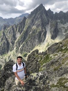 A man wearing a white shirt with colored stripes and a backpack stands on a rocky mountain ledge, smiling at the camera. Jagged, high peaks covered in grey rocks and patches of green form the rugged mountain range in the background under an overcast sky, capturing the essence of his hiking tour.