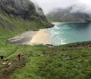 Hikers on a guided hiking tour ascend a grassy hillside towards a secluded beach surrounded by rocky cliffs. The beach features golden sand and turquoise waves gently lapping the shore. Mist hangs over the mountains, adding a mystical feel to the scenic landscape.