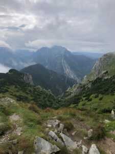 View of a mountainous landscape under a cloudy sky. The scene features rugged peaks, lush green valleys, and rocky paths. Foreground consists of a rocky trail with grass and shrubs, leading towards the distant, mist-covered mountains—perfect for an adventurous hiking tour.