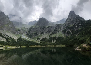 A serene mountain landscape featuring a dark, calm lake in the foreground sets the perfect stage for an unforgettable hiking tour. Rugged, green mountains with rocky peaks rise majestically under a cloudy sky, their beauty doubled as the scenery is reflected in the tranquil waters below.