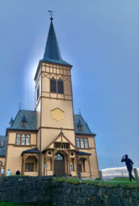 A person in black clothing stands near a historic wooden church with tall spires and intricate architectural details, likely a stop on a picturesque hiking tour. The sky is clear, and sunlight creates a halo effect around the church. Green grass and a stone wall are in the foreground.