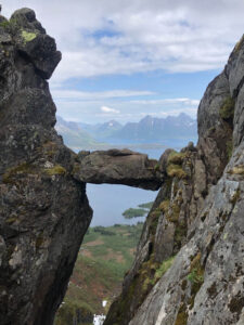 A large rock is wedged between two steep cliff faces, forming a natural bridge. Below, green vegetation and a body of water are visible, with majestic mountains in the distance under a partly cloudy sky—a perfect sight for any hiking tour.