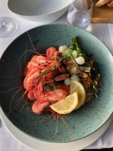 A teal speckled bowl containing fresh shrimp garnished with sprigs of dill, lemon slices, mixed greens, and pickled pearl onions evokes the simplicity of a hiking tour picnic. The bowl is set on a white tablecloth alongside empty wine glasses, a breadboard with pieces of bread, and an empty plate.