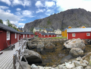 A scenic view of a coastal village with red wooden buildings along a boardwalk. In the background, there are yellow and white houses with a large rocky hill under a partly cloudy sky. Rocky terrain and patches of seaweed are visible by the water's edge, making it an ideal spot for a hiking tour.