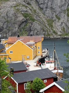 A fishing village by the water featuring red and yellow buildings, a docked fishing boat, and a rocky cliff in the background. The scene is serene with lush greenery adding some color to the landscape—a perfect stop for a scenic hiking tour.