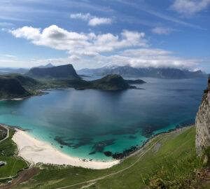 Scenic view of a pristine bay with turquoise water and a sandy beach, surrounded by lush green landscapes and rugged mountains. Perfect for a hiking tour, the sky is partly cloudy with sunlight illuminating parts of the water and mountains, creating a picturesque and serene atmosphere.