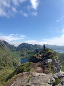 Two hikers walk along a narrow, rocky trail on a mountain ridge during their hiking tour on a clear day. The path overlooks a lush green valley, with steep mountains and a serene lake in the background under a blue sky with scattered clouds.