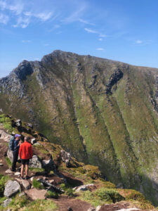 Two hikers stand at the edge of a rocky trail, looking out over a steep, green mountainside under a clear blue sky. One hiker wears a red jacket and shorts, while the other is dressed in darker clothing. The rugged, mountainous landscape suggests a challenging hiking tour ahead.