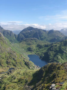 A breathtaking view of lush green mountains under a clear blue sky, with a serene lake nestled among the peaks. The landscape, ideal for a hiking tour, is dotted with patches of rocky terrain and winding paths, creating a picturesque and tranquil natural scene.