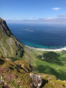 View of a coastal landscape with green fields leading to a white sandy beach, clear turquoise waters, and a deep blue ocean. A mountain slope on the left frames the scenery, perfect for a hiking tour, with small islands visible in the distance under a blue sky.