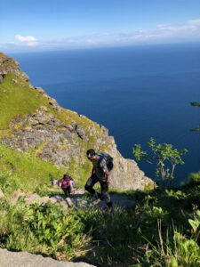 Two hikers on a hiking tour ascend a steep trail along a rocky, grassy cliffside with a vast blue ocean in the background. The sky is clear with a few clouds on the horizon. One hiker in the foreground wears a backpack, while the other follows a few steps behind. Vegetation frames the path.