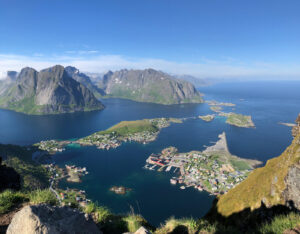 An aerial view of a picturesque coastal village nestled among rugged mountains and surrounded by clear blue water. Numerous houses and buildings line the shoreline, with rocky greeneries and peaks perfect for a hiking tour, all under a bright blue sky with a few clouds.