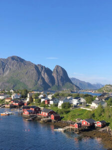 A scenic coastal village with red and white houses along the shoreline, nestled against rugged mountains under a clear blue sky. The calm blue water reflects the buildings and surrounding landscape, perfect for a hiking tour through nature's finest elements.