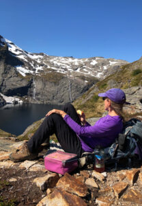 A person in a purple shirt and cap sits on a rocky ledge, enjoying an apple while overlooking a scenic mountain landscape with snow patches, steep gray cliffs, and a distant lake—a perfect moment on their hiking tour. Nearby, there's a purple lunch bag and a water bottle.