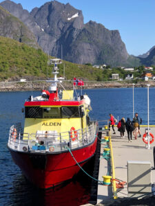 A docked bright yellow and red boat named ALDEN with people boarding or disembarking. The dock is surrounded by clear blue water, and there are towering rocky mountains with patches of greenery and white snow in the background under a clear, blue sky—perfect for a hiking tour adventure.