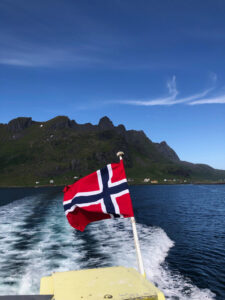 The Norwegian flag flutters at the stern of a boat as it moves through blue waters, leaving a white wake behind. Majestic, rugged mountains with sparse greenery rise in the background under a clear blue sky featuring wisps of white clouds—a perfect prelude for an upcoming hiking tour.