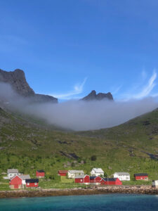 A serene coastal village with red and white houses sits beneath tall, jagged mountains. A layer of mist clings to the mountain slopes under a clear blue sky, perfect for a hiking tour. The foreground features clear, turquoise water.