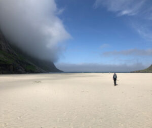 A lone person walks on a vast, empty beach with smooth sand. In the distance, misty mountains meet the ocean under a partly cloudy blue sky. To the right, a grassy hill rises towards the horizon, inviting thoughts of a hiking tour through nature's untouched beauty.