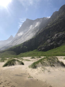 A scenic landscape showing a sunlit sandy beach with patches of grass. Behind the beach, towering rocky mountains rise under a blue sky, with wisps of clouds near the peaks. The sun illuminates the scene from the left, casting light on the mountain slopes—a perfect backdrop for an unforgettable hiking tour.