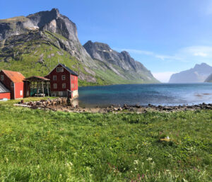 A red house with a smaller red shed sits at the edge of a calm, clear blue lake. Towering, green hills and rocky mountains loom in the background under a sunny, blue sky. The foreground is lush with green grass and wildflowers, making it an idyllic spot for a hiking tour.