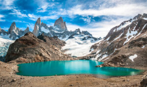 A breathtaking view of a turquoise glacial lake surrounded by rocky terrain and snow-capped peaks under a bright blue, slightly cloudy sky awaits on this extraordinary hiking tour. Towering mountains in the background include jagged, sharp summits. The setting appears remote and serene.