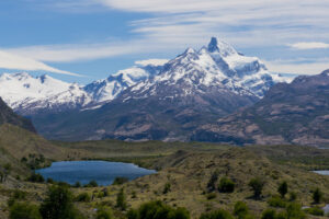 Scenic view of a mountainous landscape featuring a snow-capped peak in the background. Below the peak, there are rocky hills, lush green fields, and a small blue lake surrounded by trees. A partly cloudy sky adds depth to the picturesque scene, perfect for an unforgettable hiking tour.