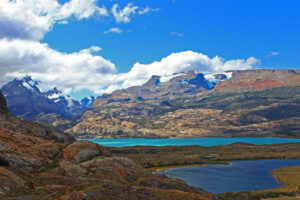 A stunning mountainous landscape with snow-capped peaks in the background, a bright blue lake in the mid-ground, and a smaller greenish lake surrounded by rocky and grassy terrain in the foreground. Perfect for a hiking tour, fluffy white clouds scatter across the blue sky.