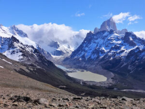 A panoramic view of a mountainous landscape featuring prominent snow-capped peaks, a large glacial lake surrounded by rugged rocks and sparse greenery, and a blue sky with scattered clouds. The terrain is rocky in the foreground, leading to the serene lake in the distance—a perfect setting for a hiking tour.