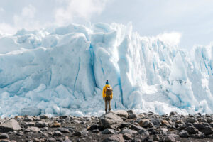 A person in a yellow jacket and green hat stands on a rocky terrain during a hiking tour, facing a massive ice formation. The towering ice appears blue and white, showcasing complex layers and textures. The sky above is partly cloudy.