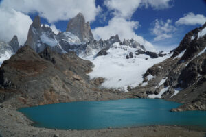 A turquoise glacial lake is nestled among rugged, rocky terrain with snow-capped peaks and jagged mountain ridges in the background. The sky is blue with scattered clouds, making it an ideal spot for a hiking tour that captures views of natural beauty and serenity.