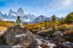 Person wearing a green jacket and hat sits on a large rock, enjoying a scenic hiking tour with a flowing stream, colorful autumn foliage, and towering snow-capped mountains under a clear blue sky.