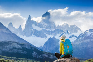 A person with a blue backpack wearing outdoor gear sits on a rock, gazing at a breathtaking mountain range with snowy peaks and low clouds. The vibrant scene captures the essence of a hiking tour, blending green valleys and rugged, majestic mountains in the background.